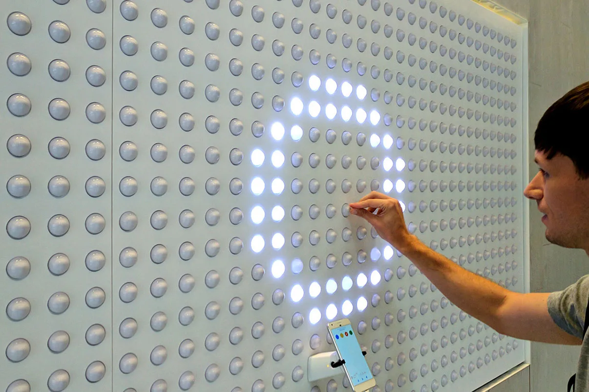 Close up of a hand drawing a bright shape into the grid of white dots, a phone docked on the wall below