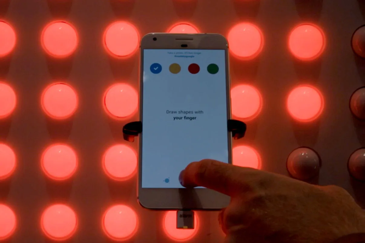 A Google Pixel phone held against red-lit dots, the app on screen prompting you to draw shapes with your finger