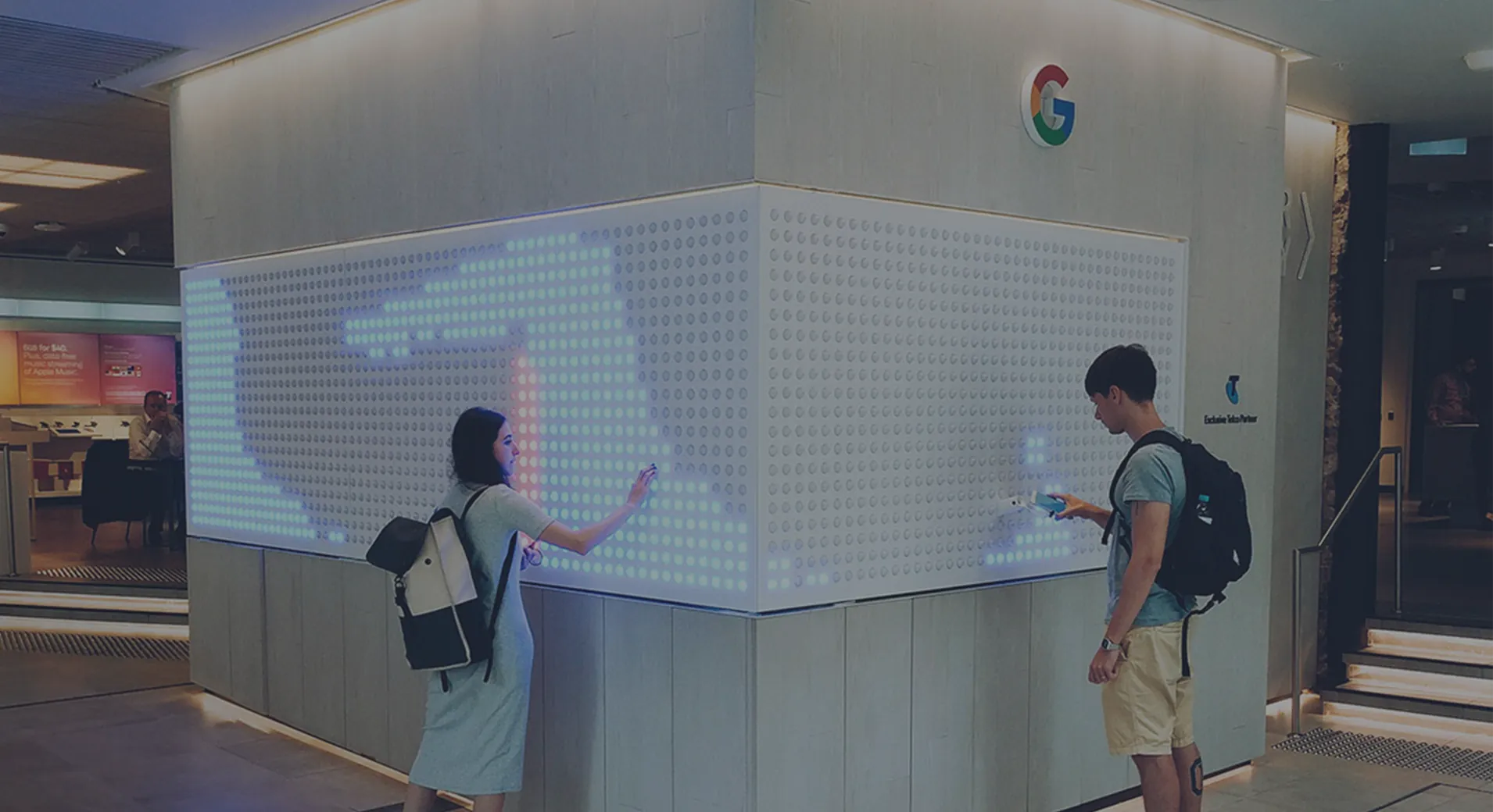 Two shoppers tracing shapes on the Google Pixel wall in a Telstra store, its grid of dots lit pale blue around the corner