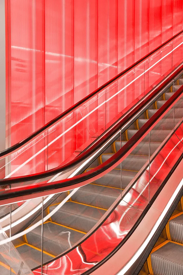 Close view of the escalator handrails against the red wall, the glass balustrade catching the glow