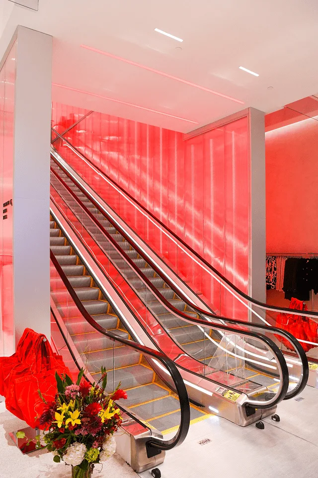 The foot of the escalator with a flower arrangement and red fabric drape, the lit wall rising behind