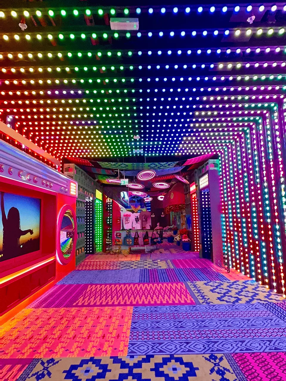 Passage with the ceiling and walls covered in rainbow pixel dots, screens down one side and a patterned floor running away