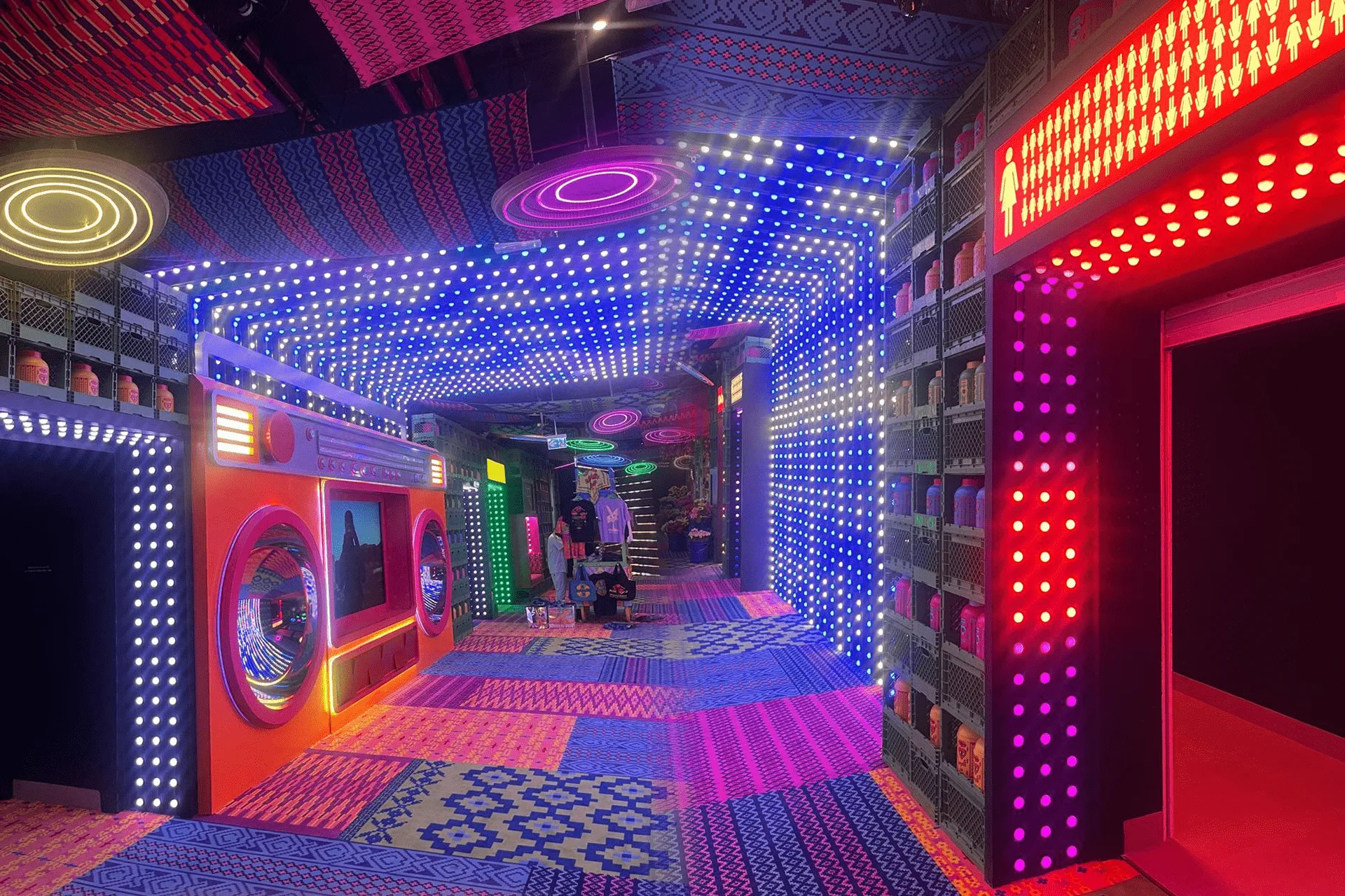 Corridor at House of Hype lined with pixel dot walls in blue and magenta, a red neon doorway on the right and patterned carpet underfoot