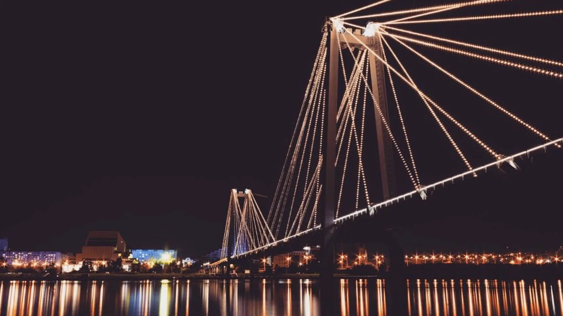 The bridge in warm amber and white at night, city lights and long reflections on the water beneath the span