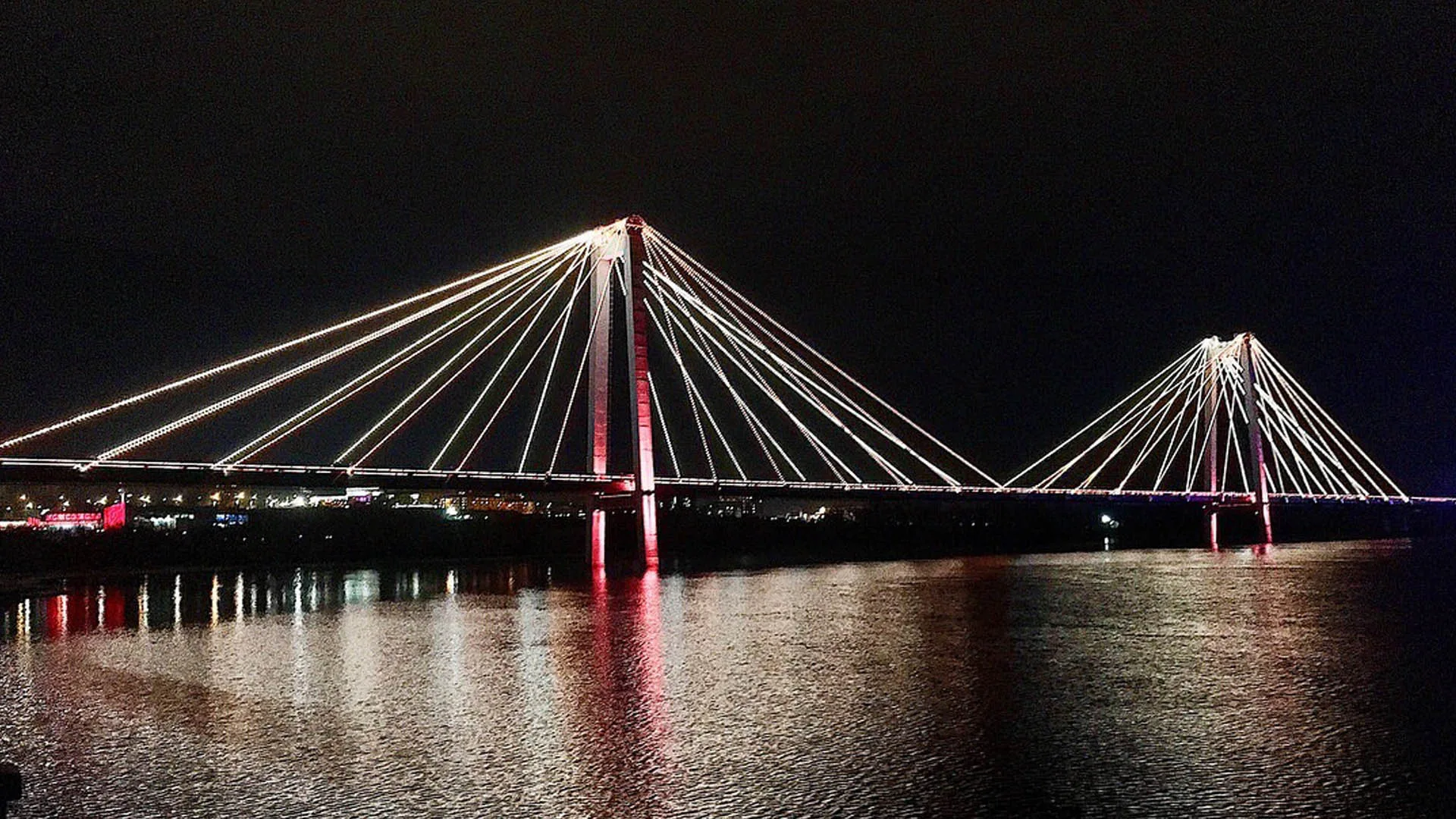 Seen from the opposite bank, cables picked out in cool white with a red wash at the near tower and still water below