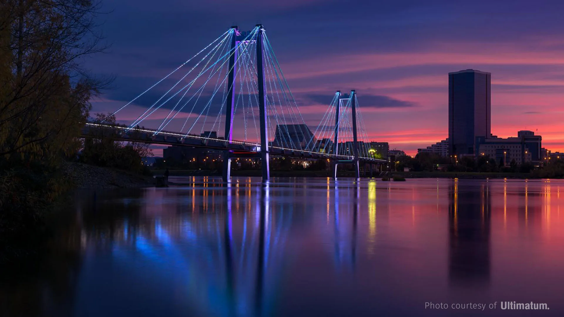 Blue-lit cables against a pink and violet sunset, a dark high-rise on the skyline and colour spilling into the river