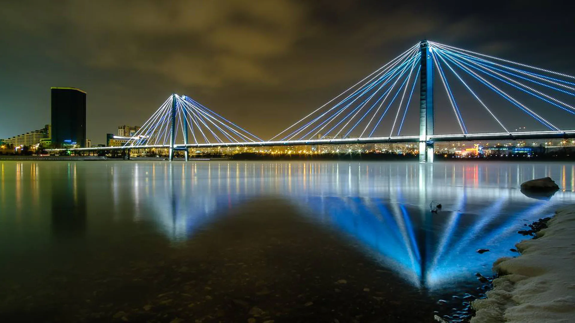 The span in deep blue from a rocky shoreline, its reflection stretching across flat water under olive cloud