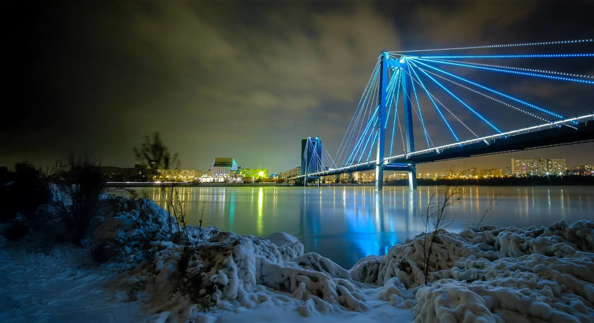 The Vinogradovsky footbridge at night from deck level, handrails glowing blue and orange with lit cables rising to the pylon