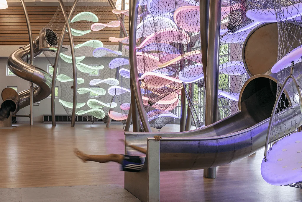 Pastel pink and green platforms glowing indoors, a child's legs dangling from the polished steel slide exit