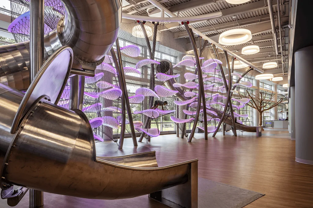 Close on the steel slide chute curving down to the floor, lilac platforms and ceiling ring lights beyond