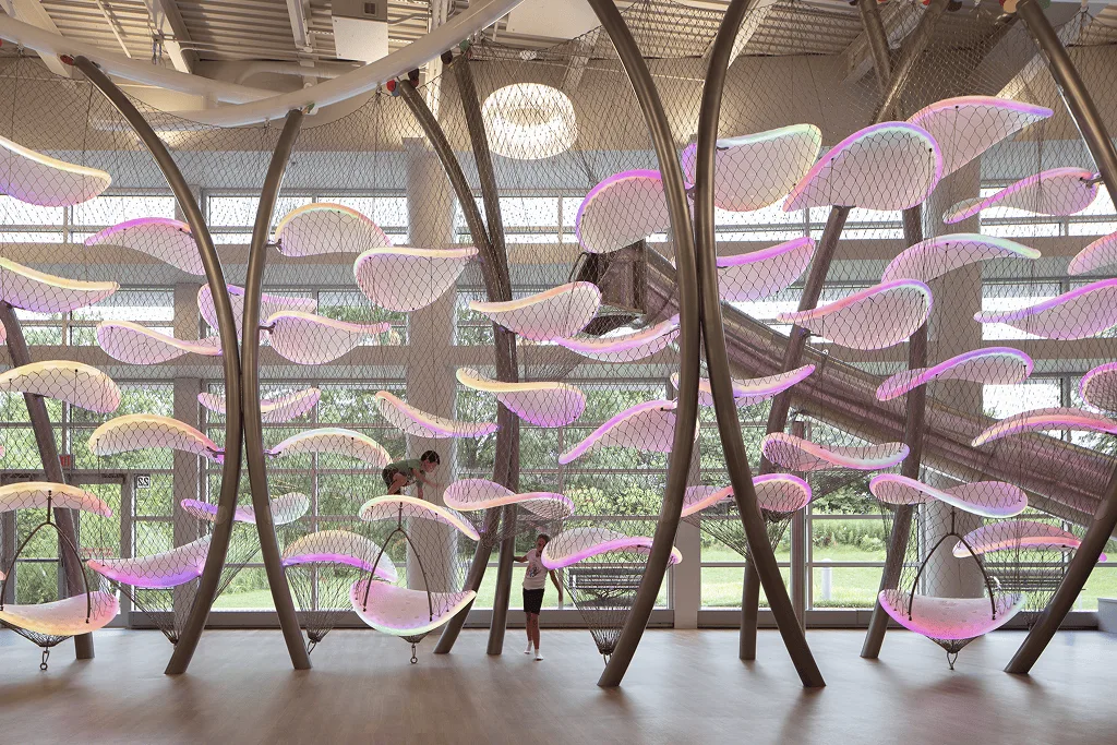 Floor level view of the climber lit pink, curved steel ribs rising past a child resting in a net hammock