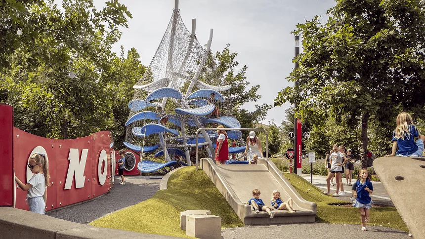 Children on a grass mound and slide outdoors, the blue and white climber rising behind a red wall of oversized letters