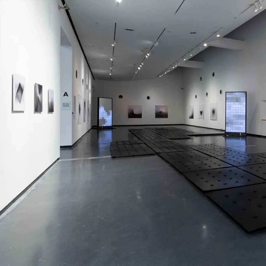 The exhibition seen down its length, prints along both walls and the dark studded floor plates leading toward the glowing monolith