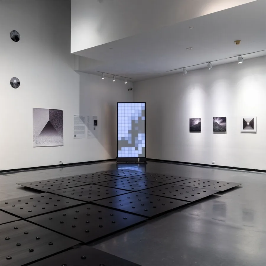 Corner of the gallery with the monolith lit between wall prints, dark floor plates and their black dots filling the foreground