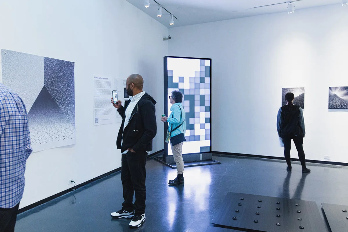 Visitors in the gallery, one photographing a print on his phone while another stands in front of the pixel monolith