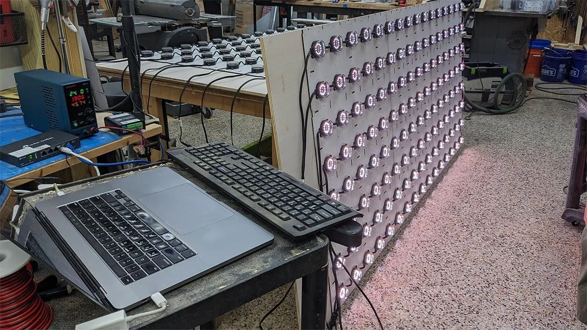 A laptop and keyboard on the workbench beside the lit panel, test patterns running across its dots