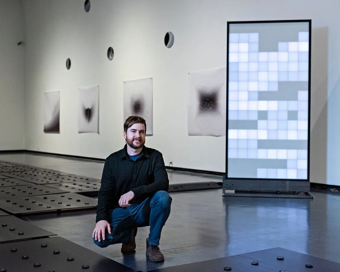 The artist in his exhibition, cropped closer, the grid of white and grey pixels glowing at his shoulder