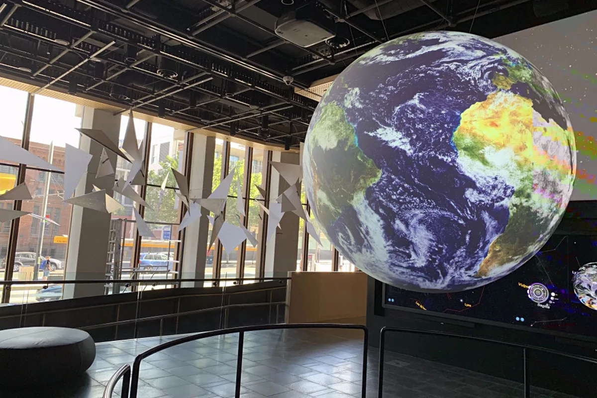 Wide view along the balcony, panels hanging overhead and the globe screen throwing light onto the concrete columns