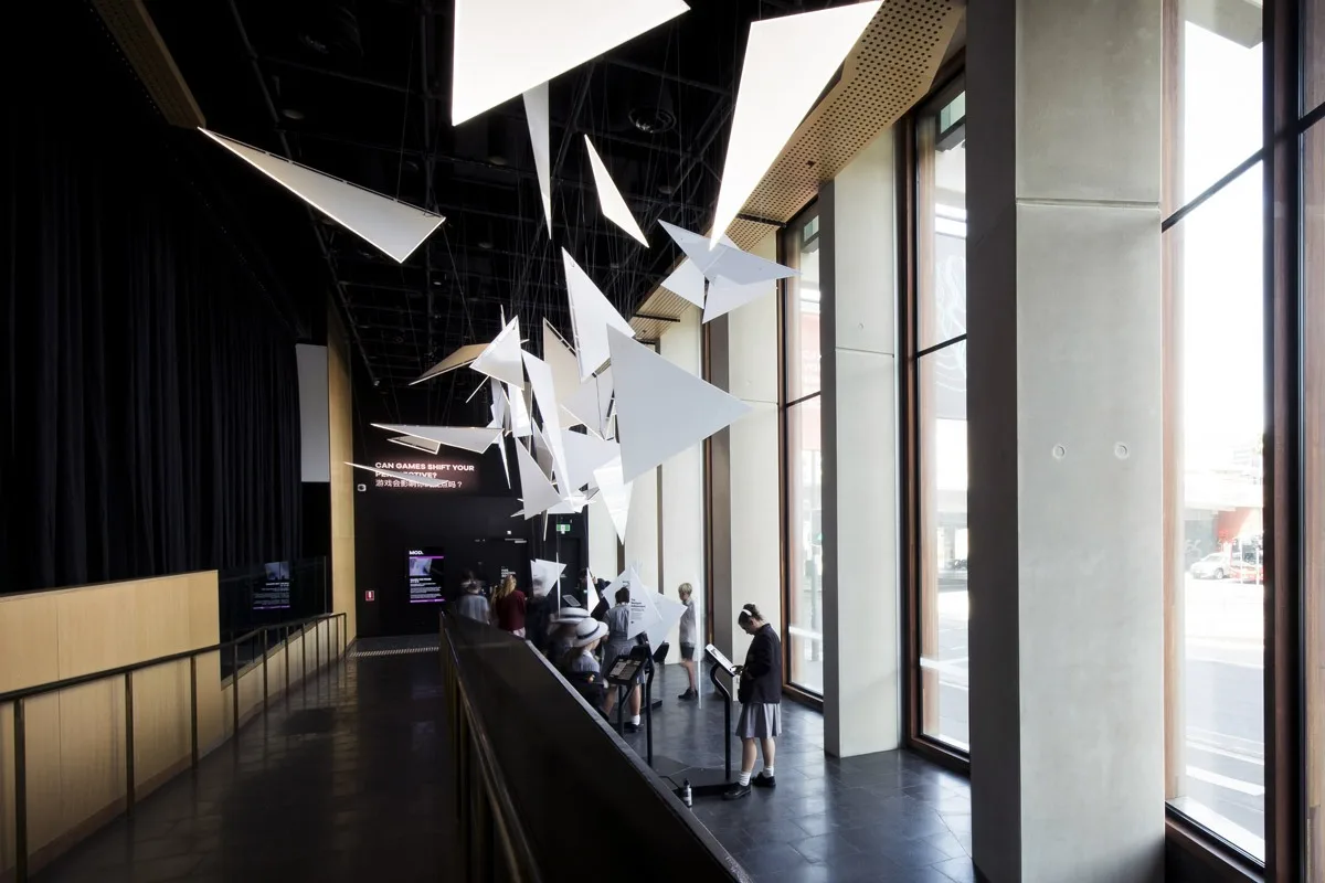 The gallery walkway from a step further back, triangles scattered overhead and visitors along the window rail in daylight