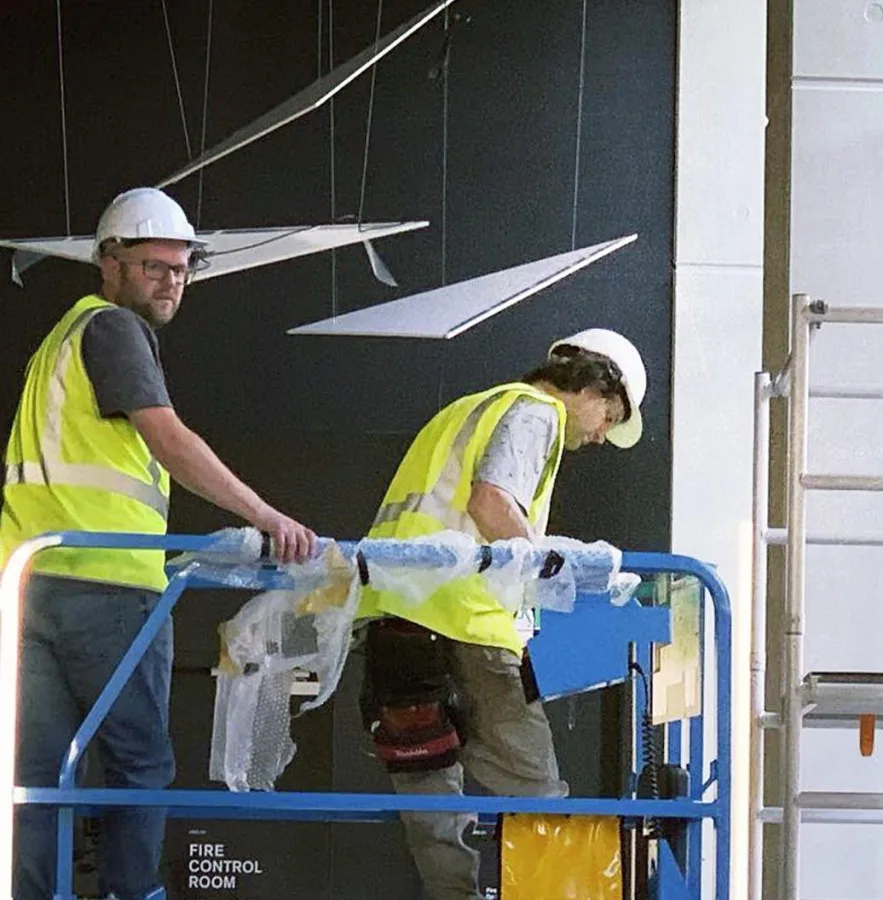 Closer on the two installers unwrapping a panel on the lift platform, the Fire Control Room door behind them
