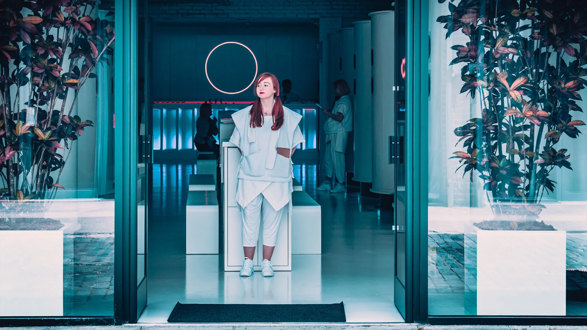 A woman in white standing in the open entrance doorway between two planters, the lit reception behind her
