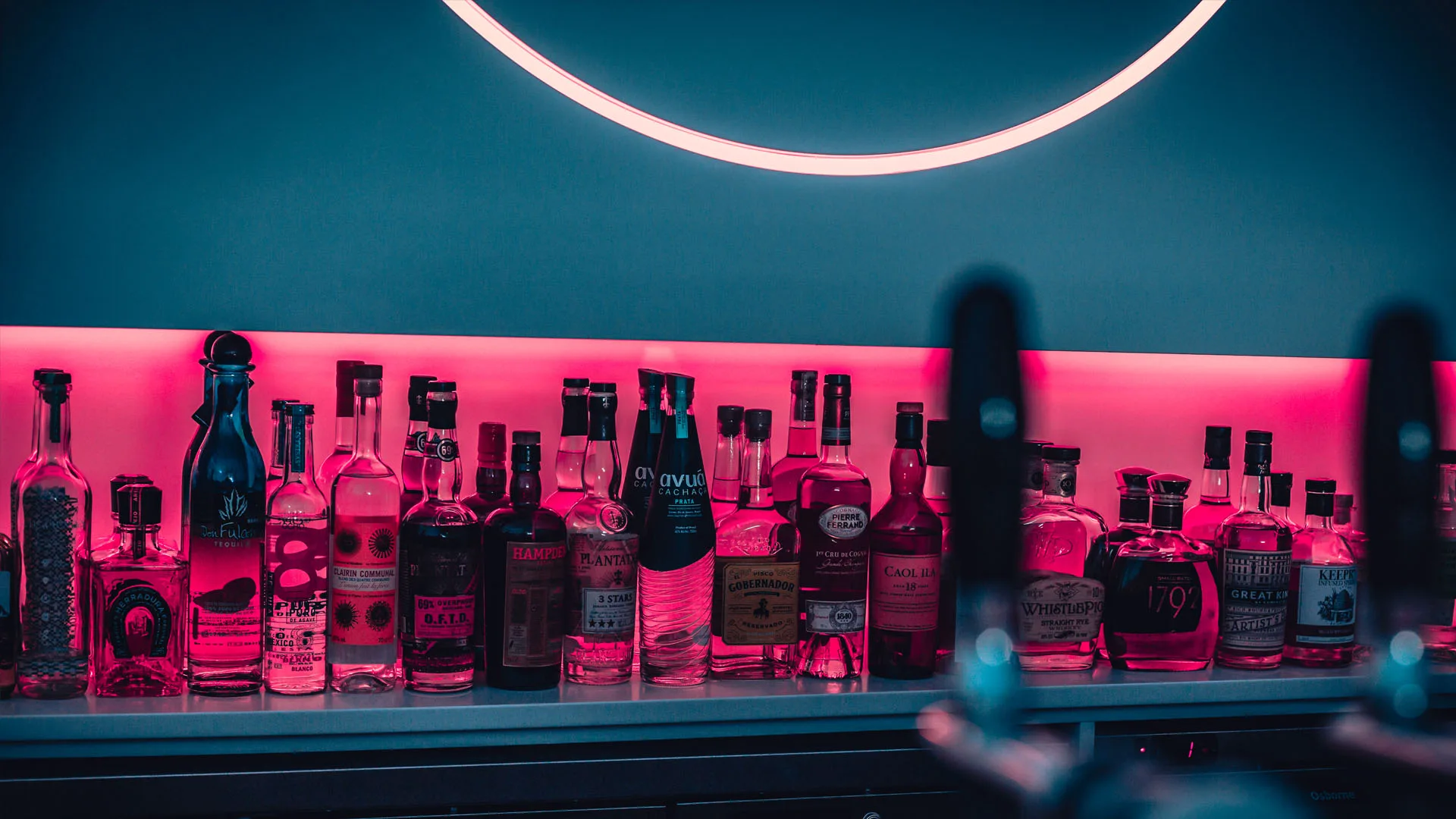Spirit bottles ranged along the back bar under hot pink backlight, a white neon ring hanging above them