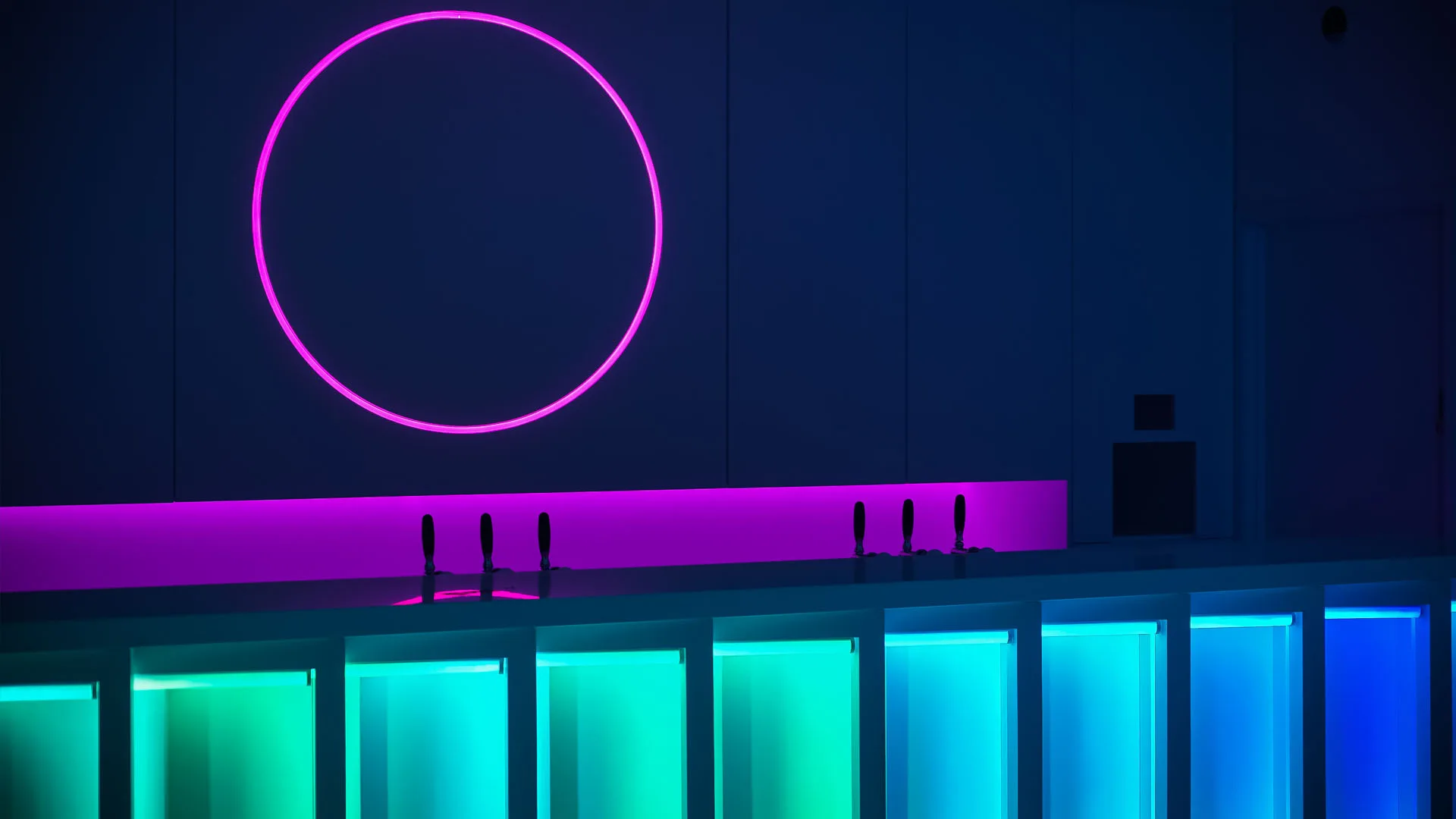 Magenta neon circle over a dark bar, the counter front glowing in bands of green and turquoise