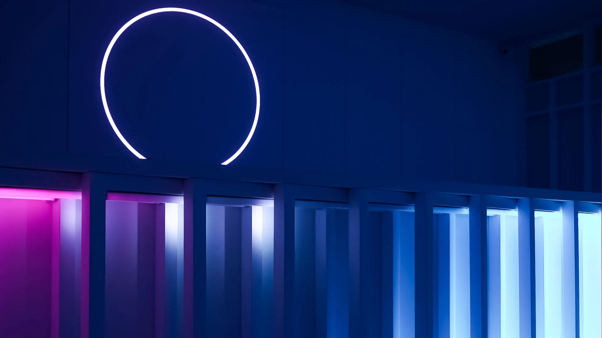 White neon ring on a blue wall above a run of tall light panels grading from magenta into pale blue
