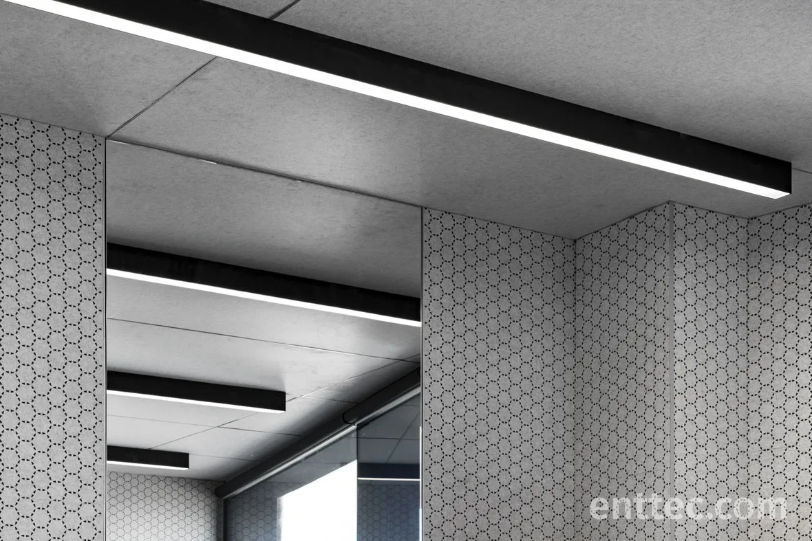 Classroom ceiling corner where a black recessed channel carries a linear luminaire, hexagon patterned acoustic panels on the walls