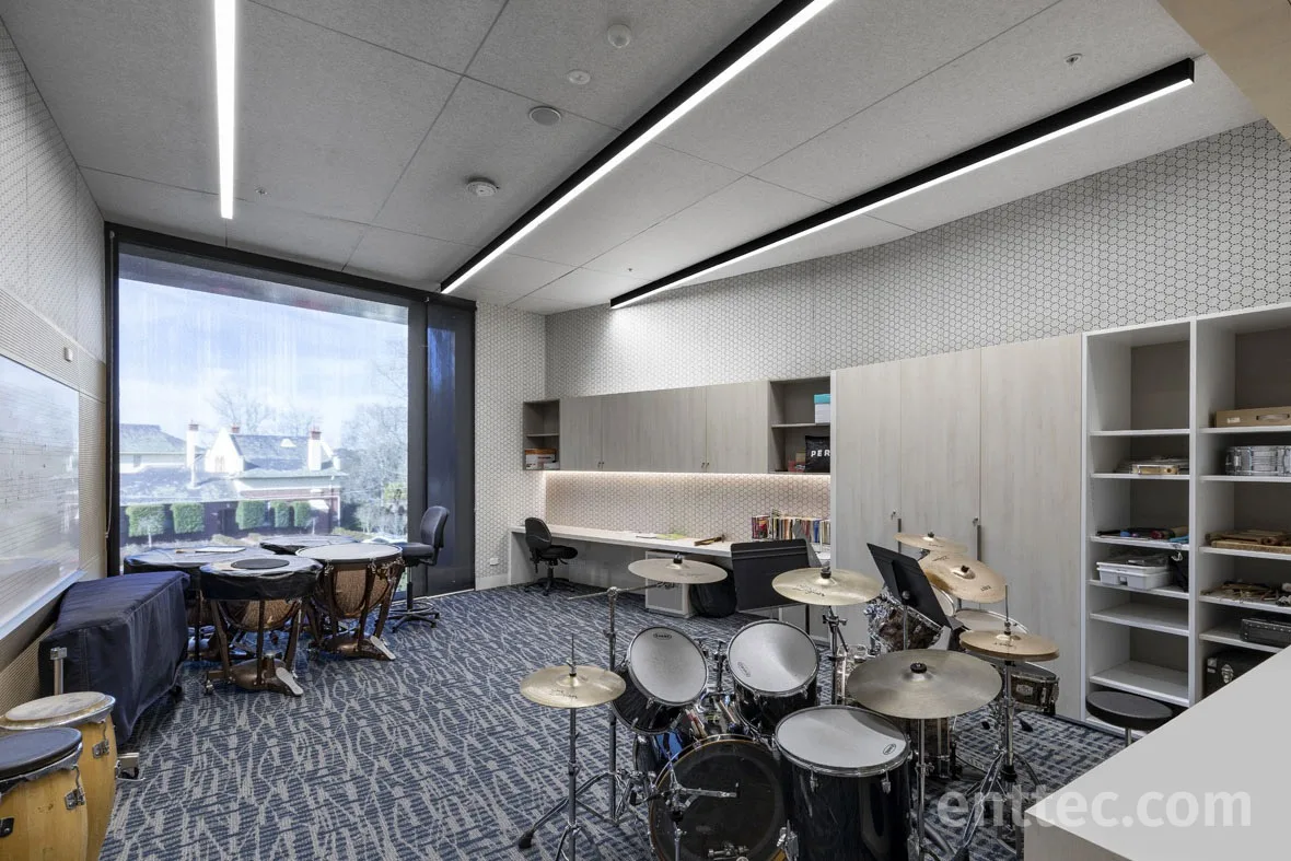 Rehearsal room with timpani and a drum kit, angled linear luminaires overhead and a lit shelf run along the back wall
