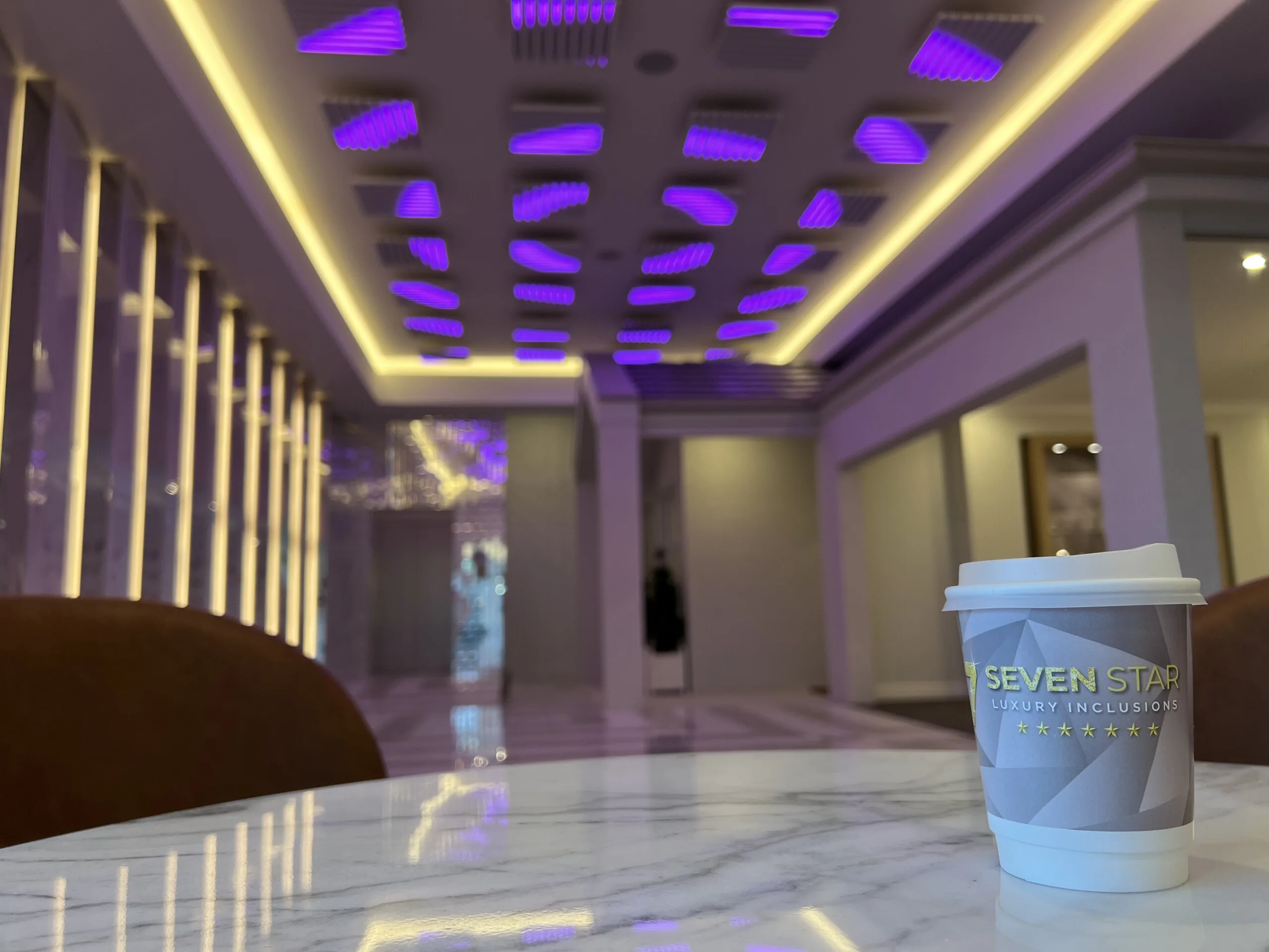 The lobby lit violet and gold, ceiling pods reflected in a polished marble counter with a Seven Star branded coffee cup in the foreground