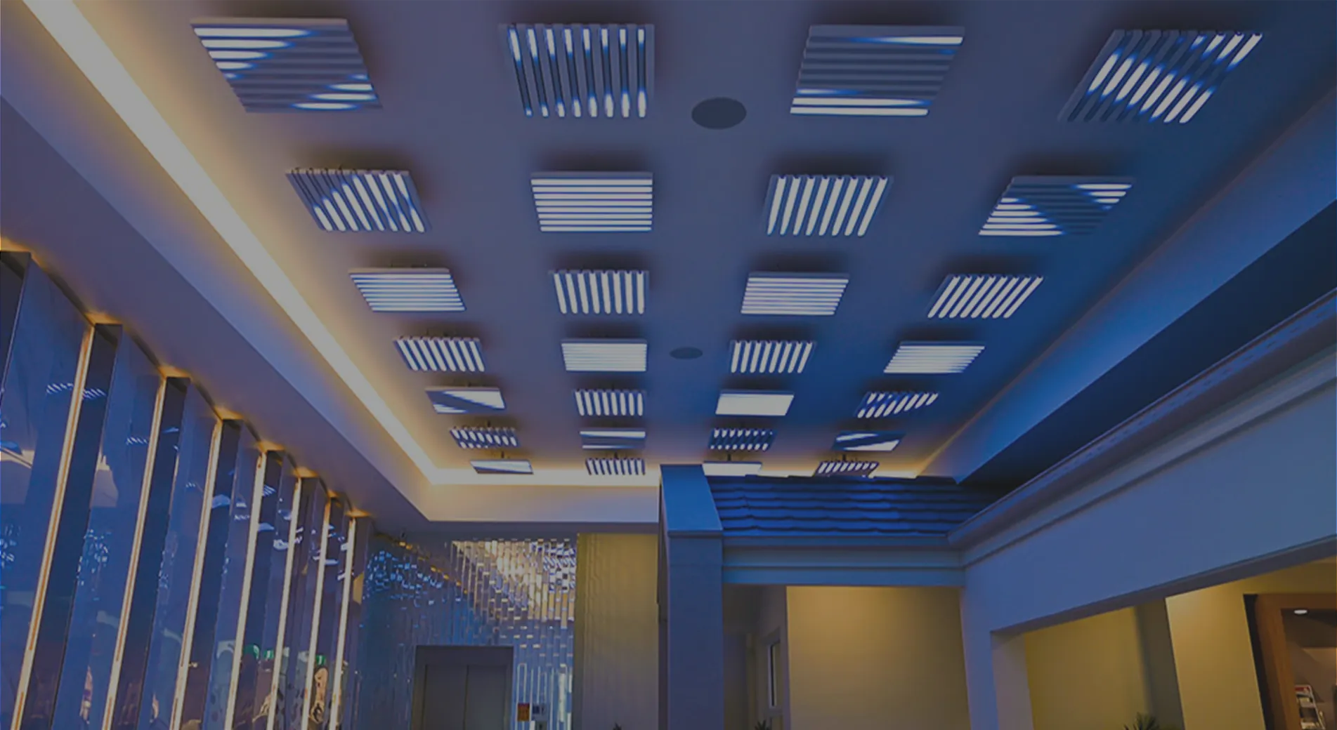 The Kaplan Homes lobby washed in deep blue, the full grid of square ceiling pods overhead with warm light spilling from the shopfronts below