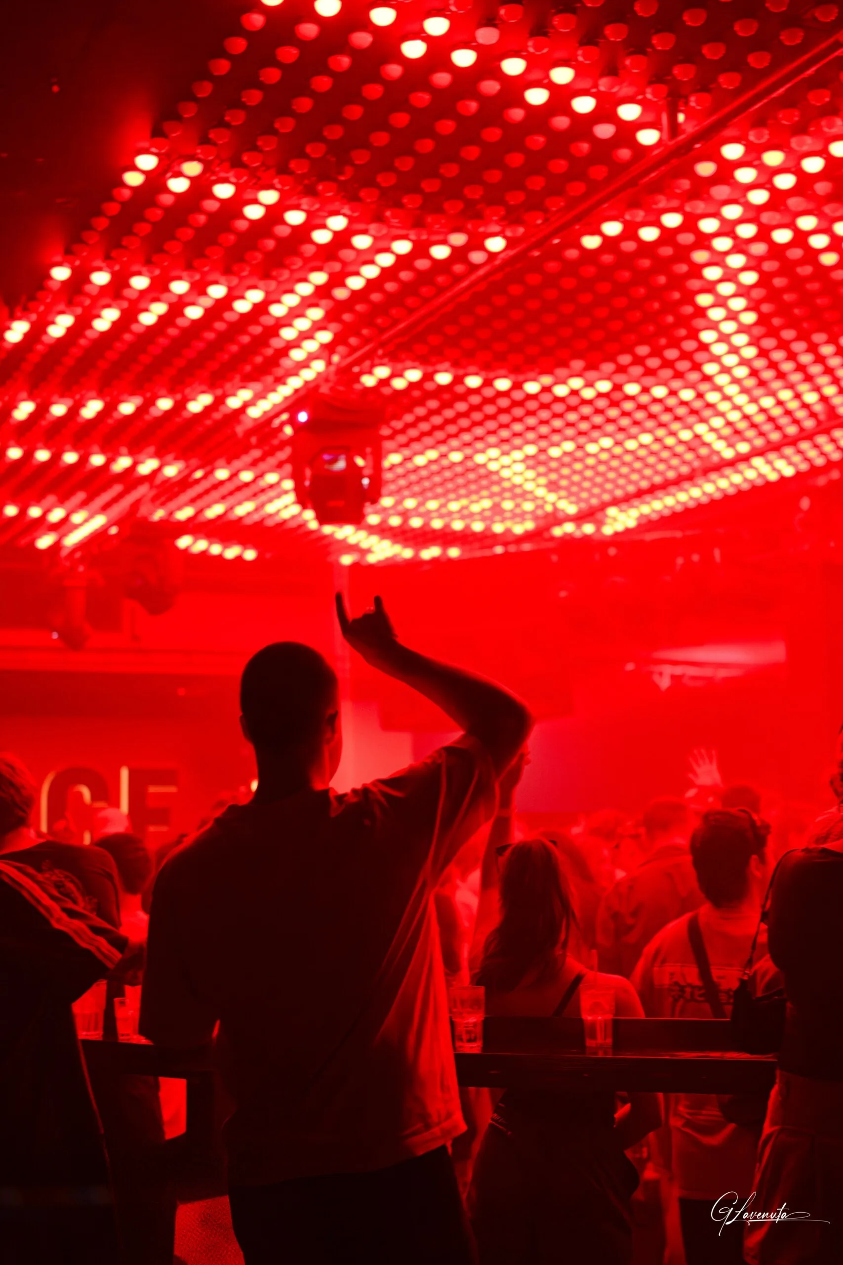 A man in a white t-shirt raising one arm under a ceiling of red dots, the room thick with red haze
