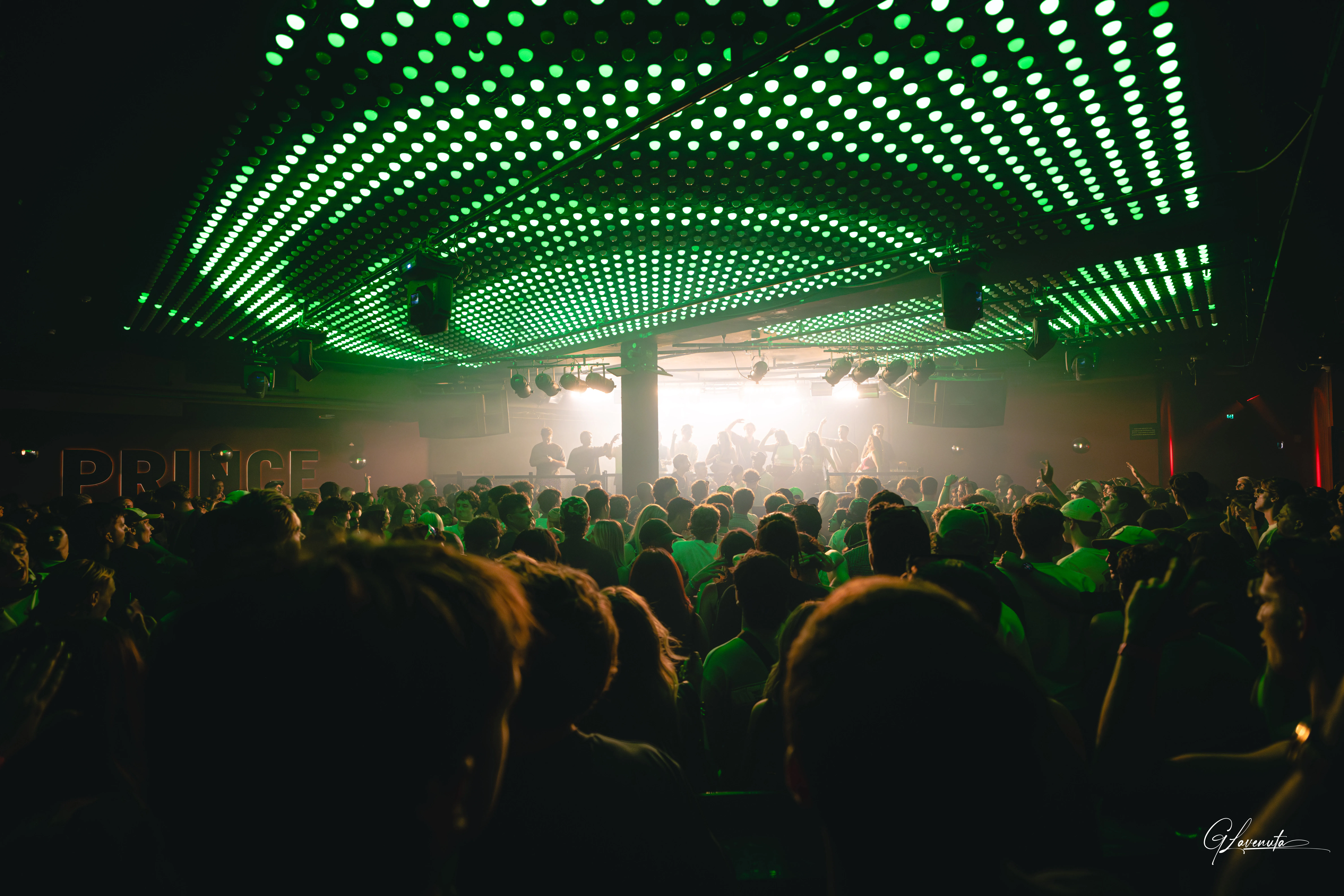 Green dot patterns running across the ceiling seen from the back of the room, the stage lit white at the far end
