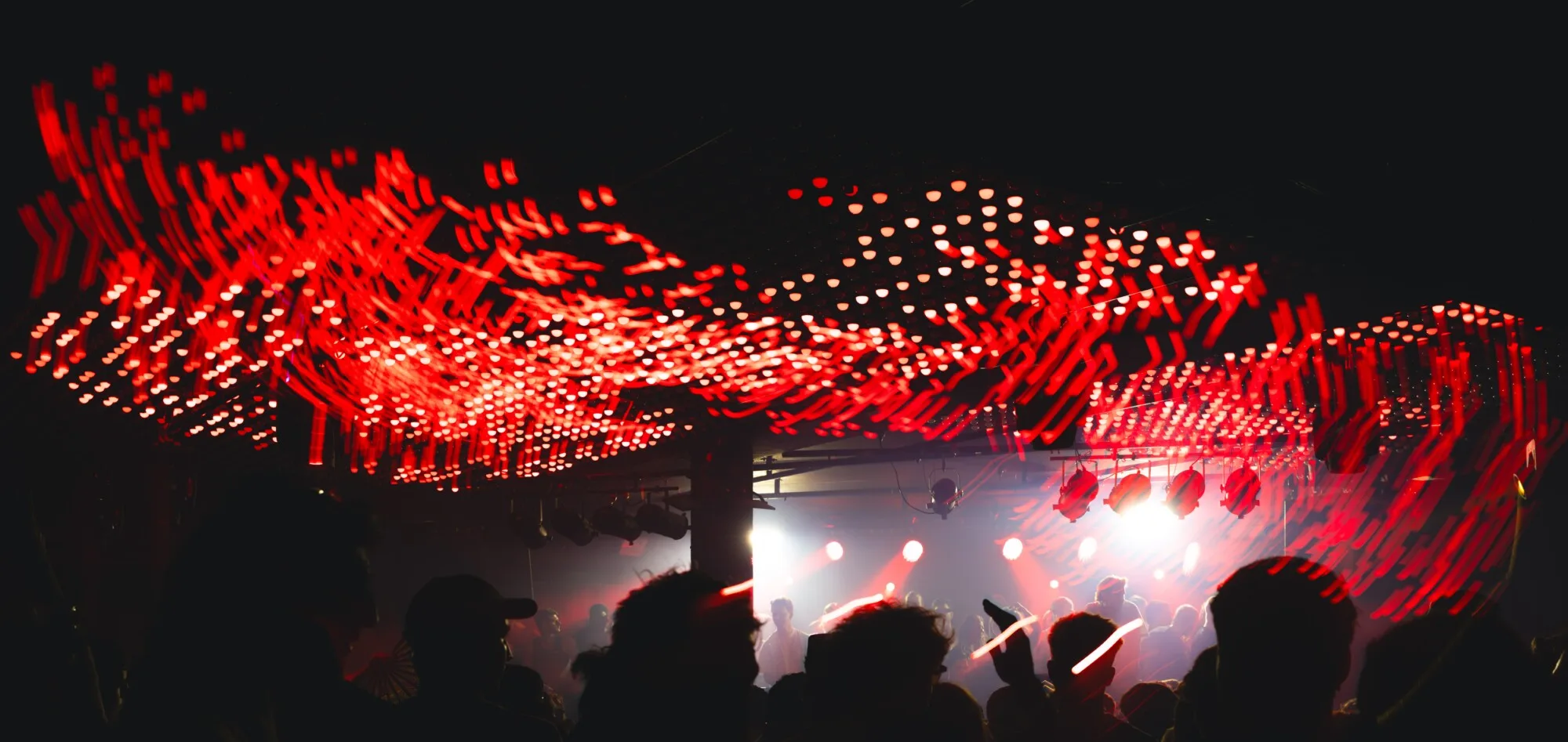 Long exposure of the red ceiling dots streaking above the crowd, the stage a bright white blur at the end of the room