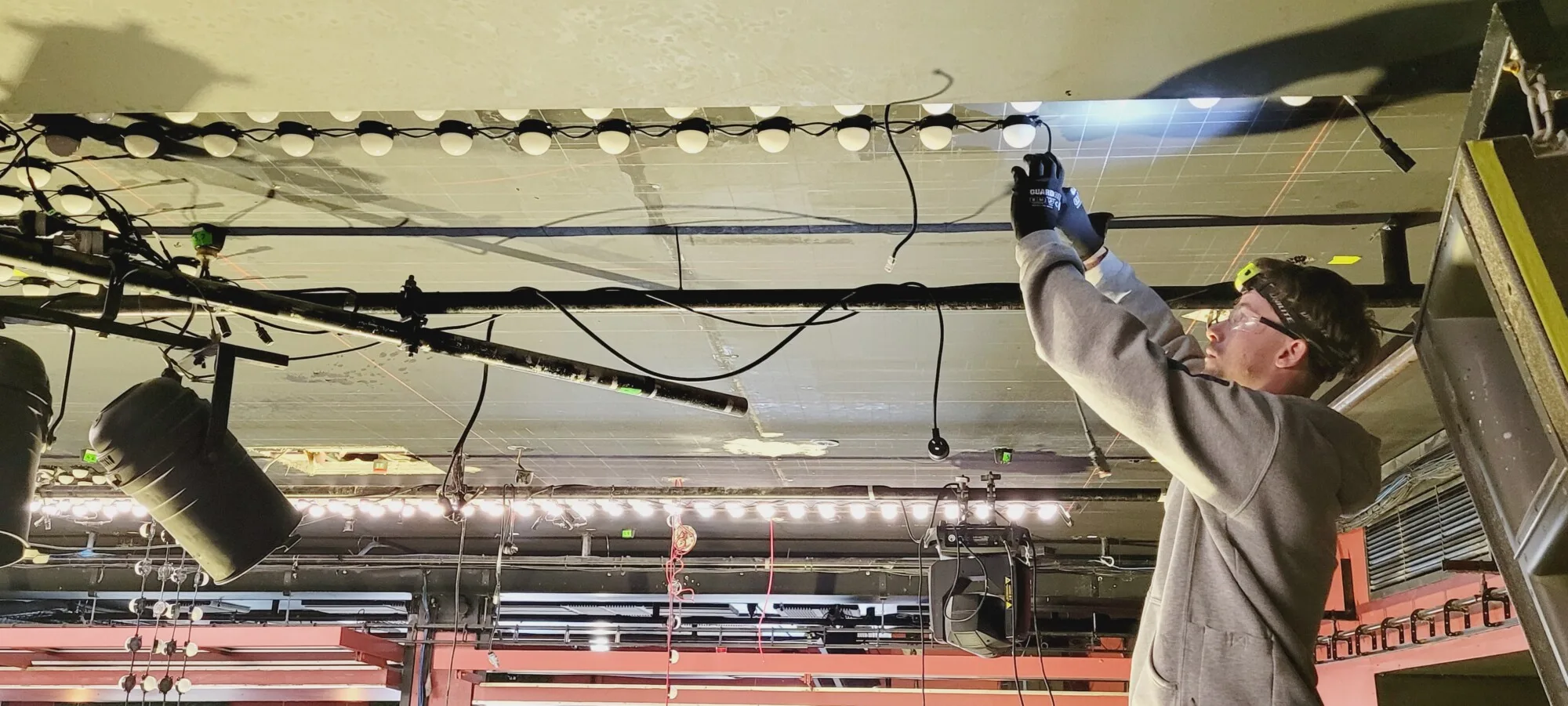 An electrician on a lift fitting a run of dots to the ceiling under work lights, cable and truss around him