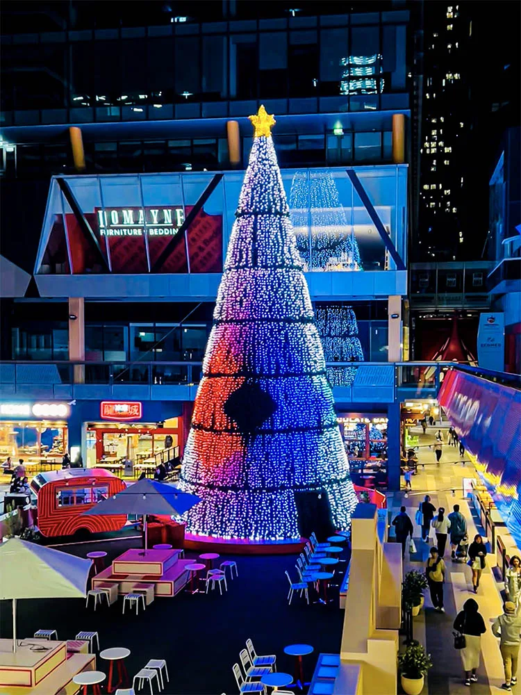 The tree from the upper level, its cone running green, blue and white while people move between the food stalls below