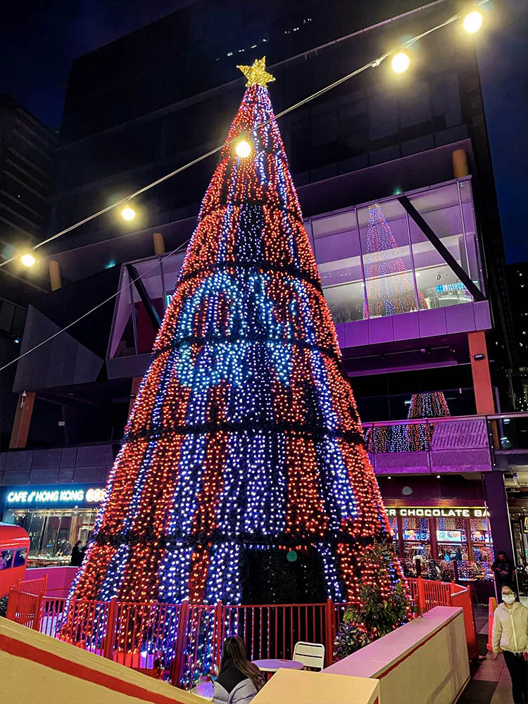 Looking up at the tree at night in red and blue, festoon lights strung overhead and the surrounding shopfronts washed in purple