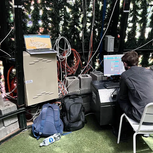 A programmer at a temporary desk behind the tree, two monitors and a laptop set up beside a control cabinet and coiled cabling