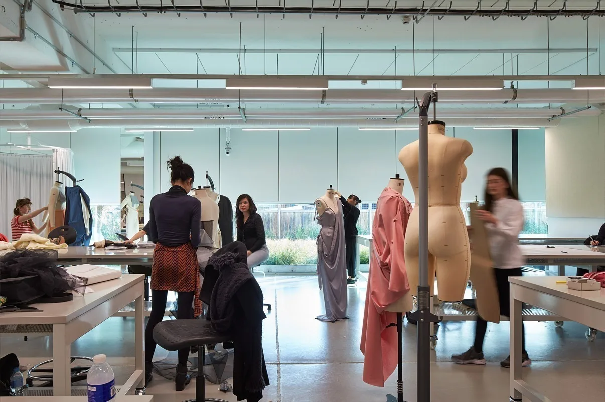 The same studio from further back, students cutting and pinning at long white tables with racks of garments behind them