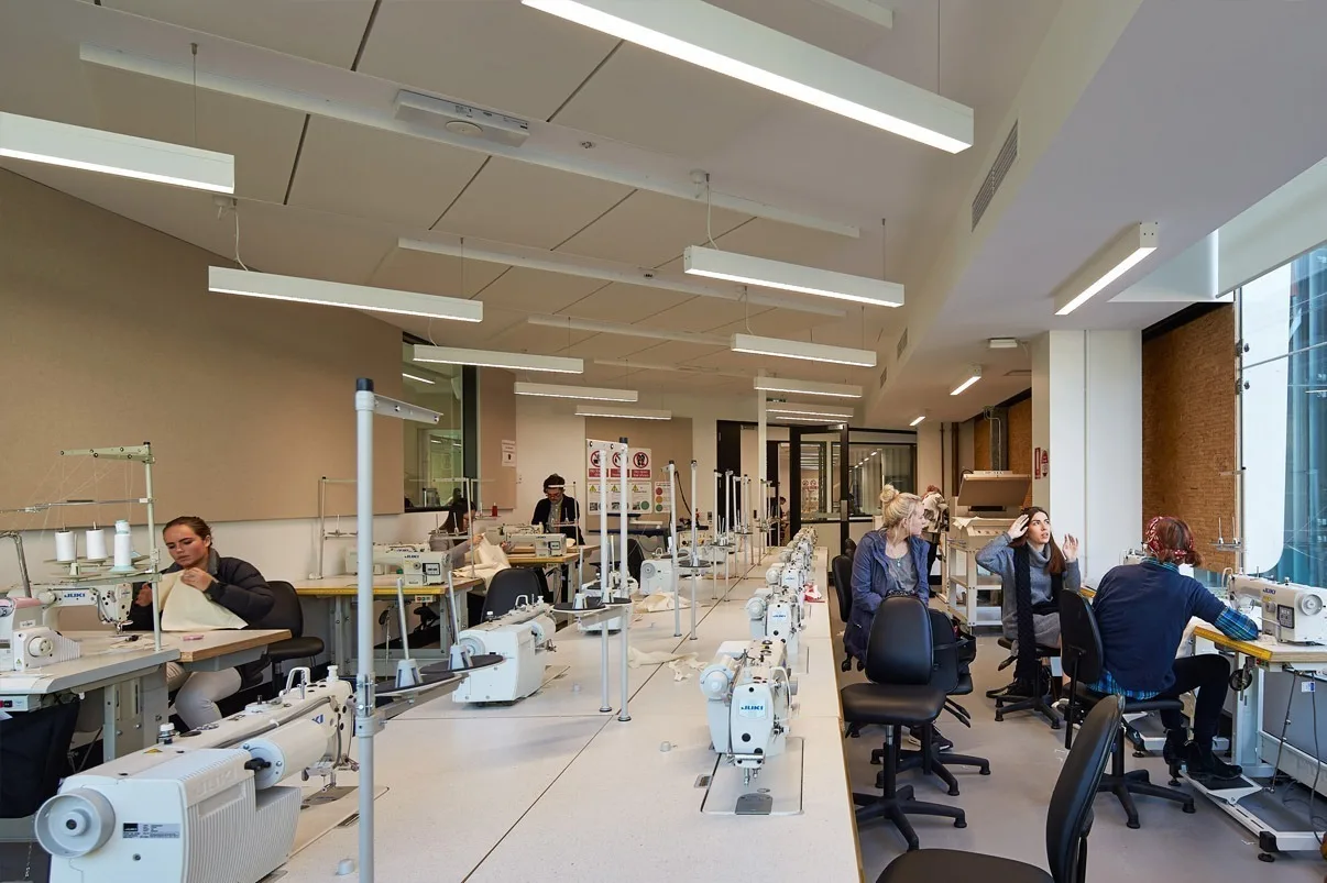 Rows of sewing machines down a long teaching room, students seated at the benches under continuous linear luminaires