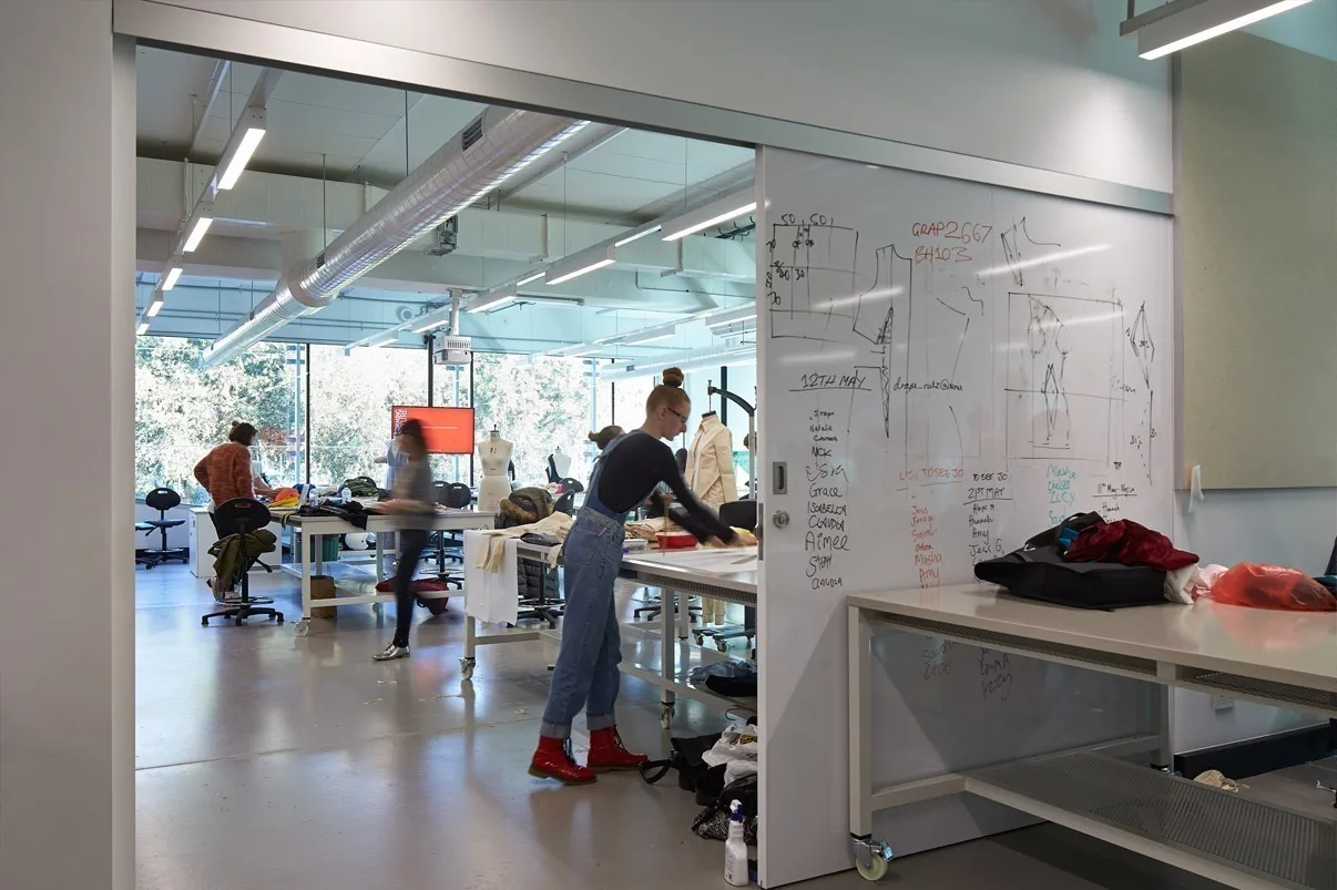 A studio seen through a glass wall, sketches and notes covering the whiteboard while students work at benches inside