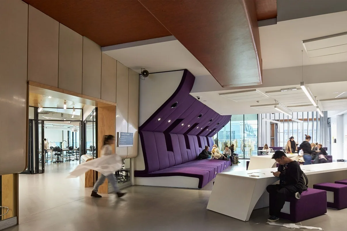 Breakout area with a sculptural purple tiered bench under a timber ceiling, a student walking past low study tables