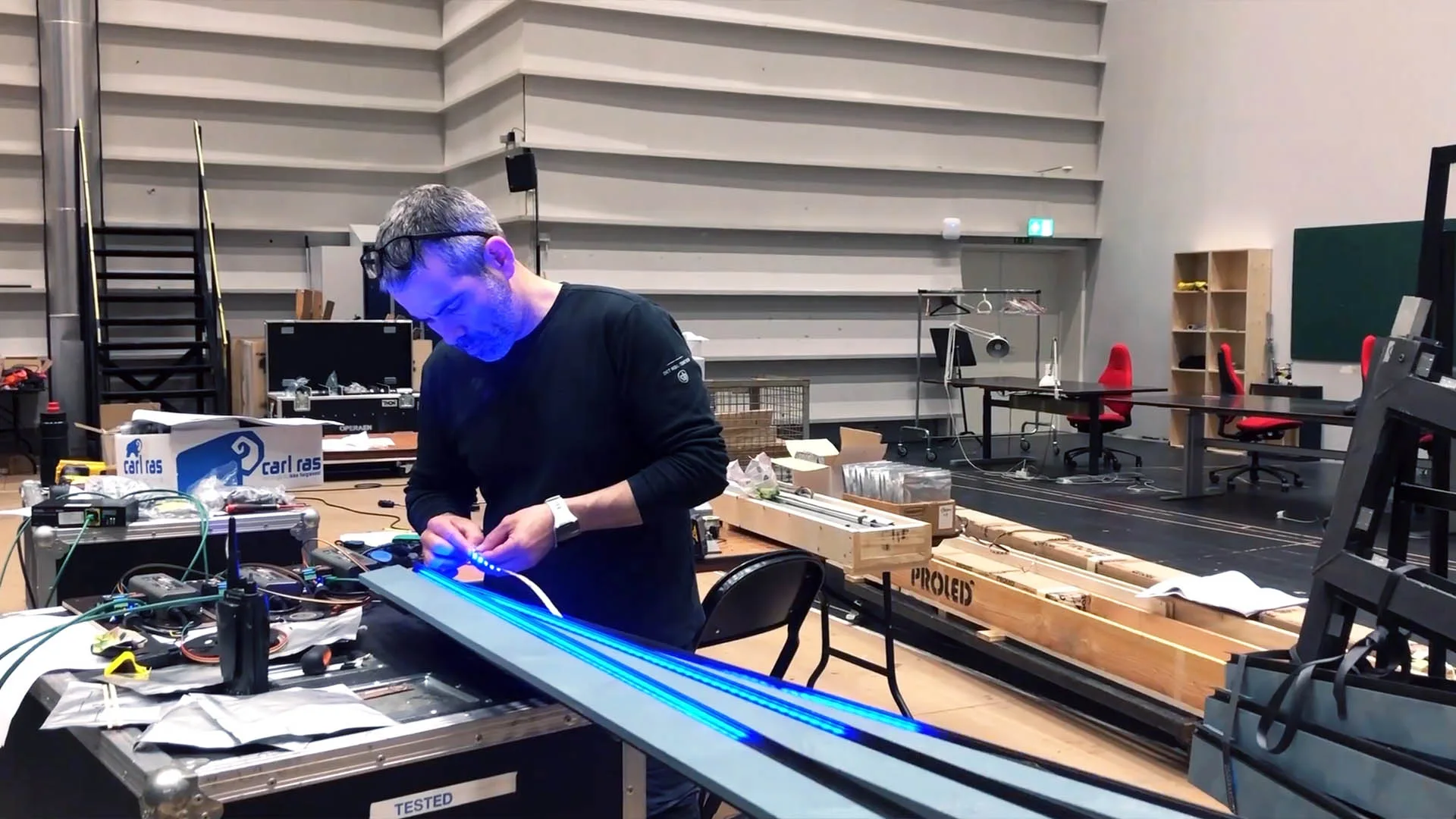 A technician at a workshop bench soldering a lit blue LED strip, his face lit by the pixels, magnifier headband on