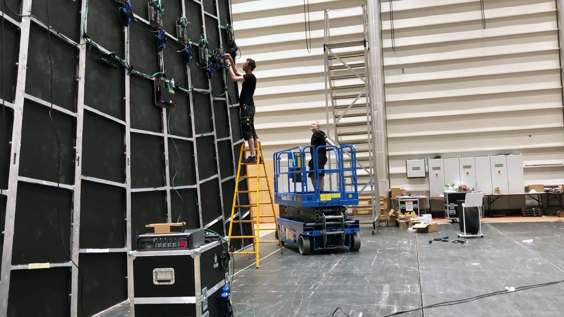 Crew on a scissor lift and a ladder fitting LED battens to a tall black grid frame in the rehearsal hall