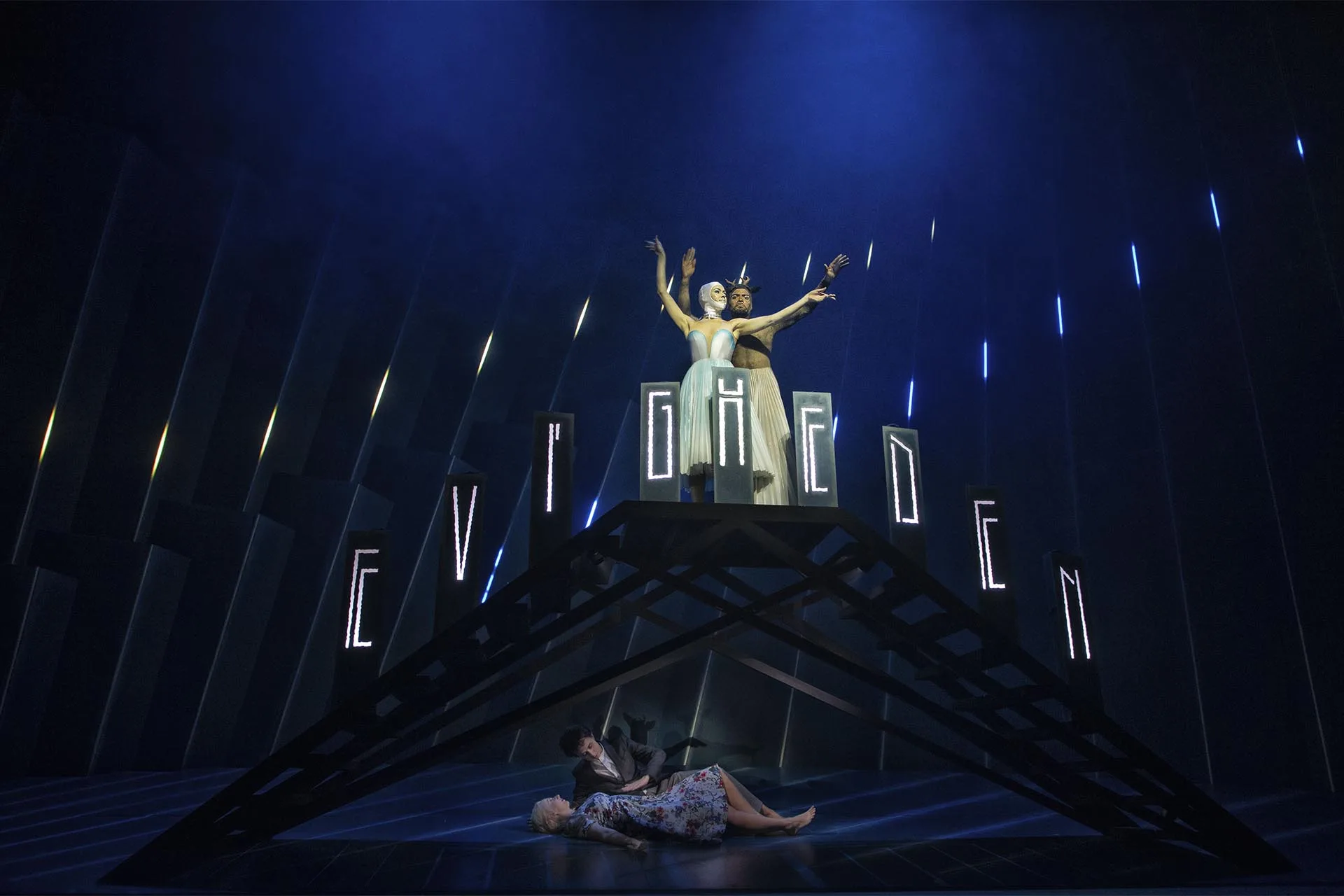 A gold-costumed singer with arms raised above a black gable, the word EVIGHEDEN spelled in light and blue pixel strips behind