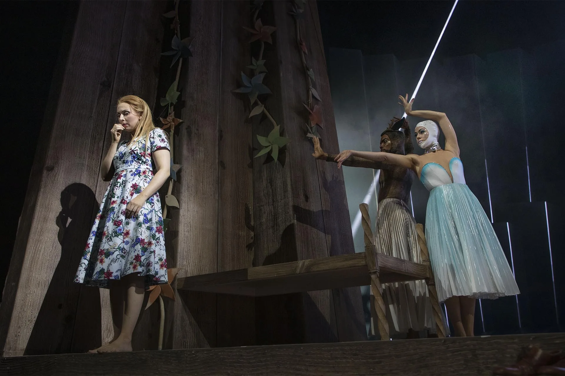 Two performers on the timber set among paper stars, one in a floral dress, one in a pale tutu with a raised blade