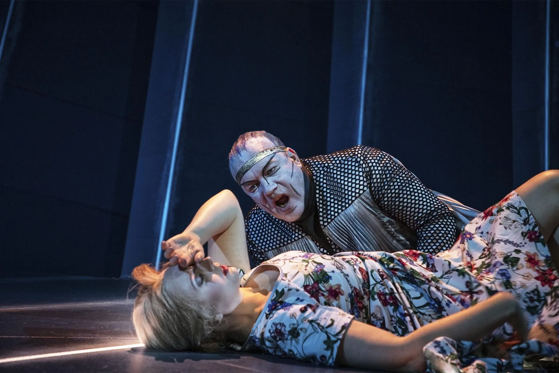 A white-faced performer leaning over a woman lying on the stage floor, thin blue pixel lines behind them