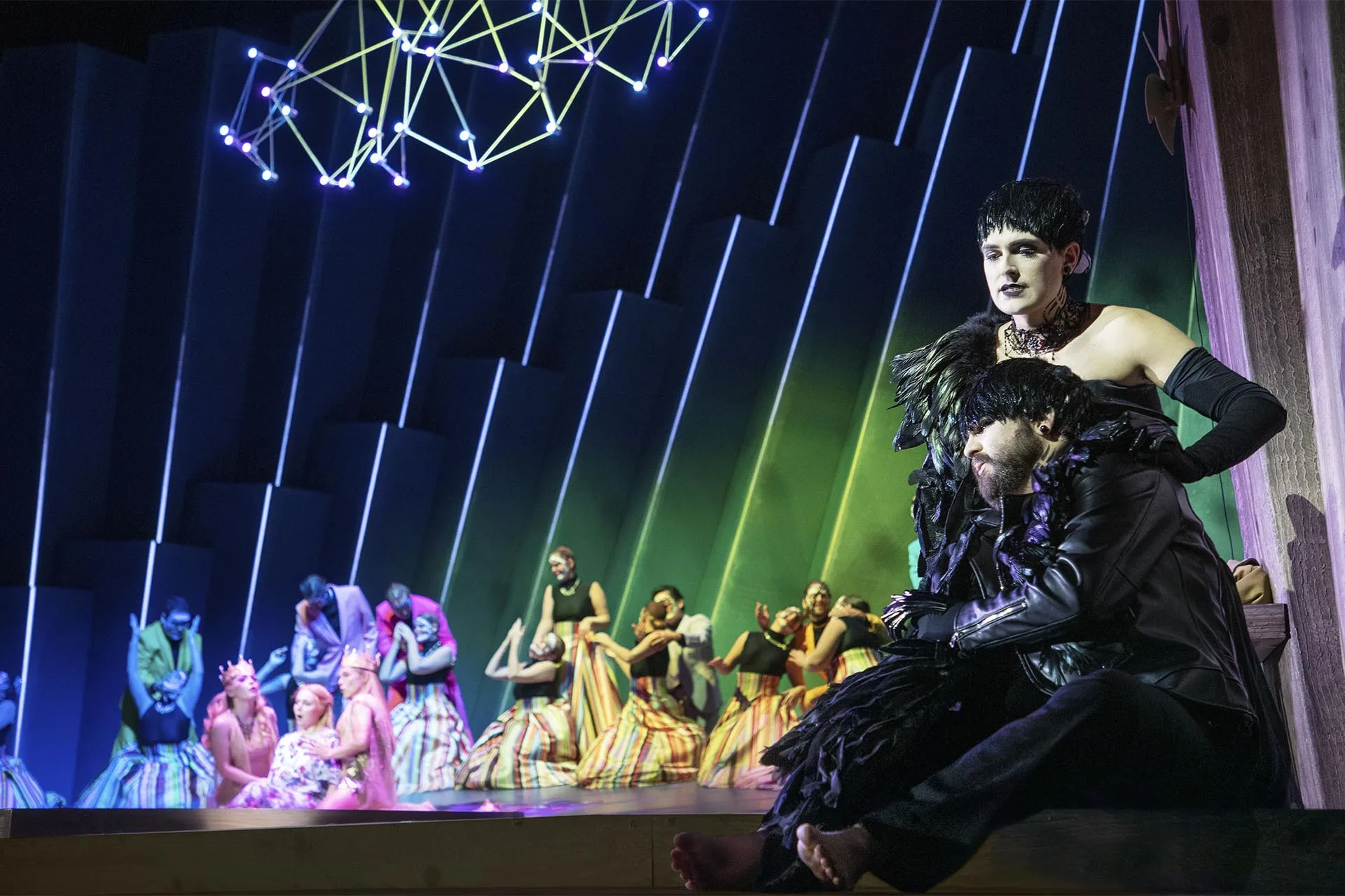 A dark-costumed figure cradling another downstage, the pastel-dressed chorus behind under green light and a cluster of pinpoint stars