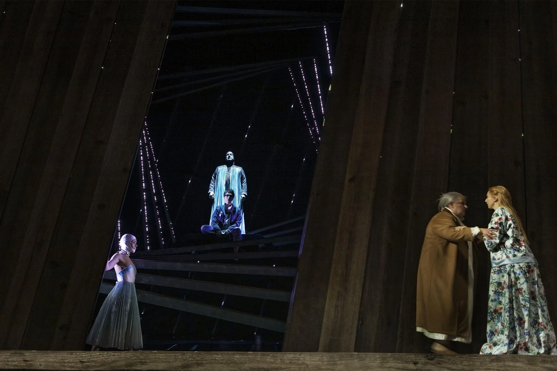 Wide dark stage with a lone singer high in a lit frame, two performers talking downstage right, strings of tiny lights falling